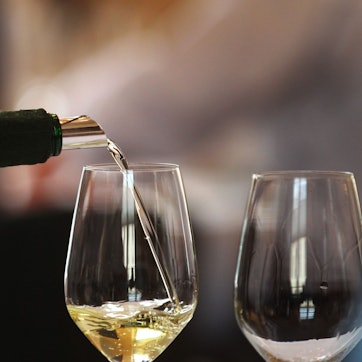 Aus einer verhüllten Flasche wird Weißwein in eins von zwei leeren Gläsern gegossen.