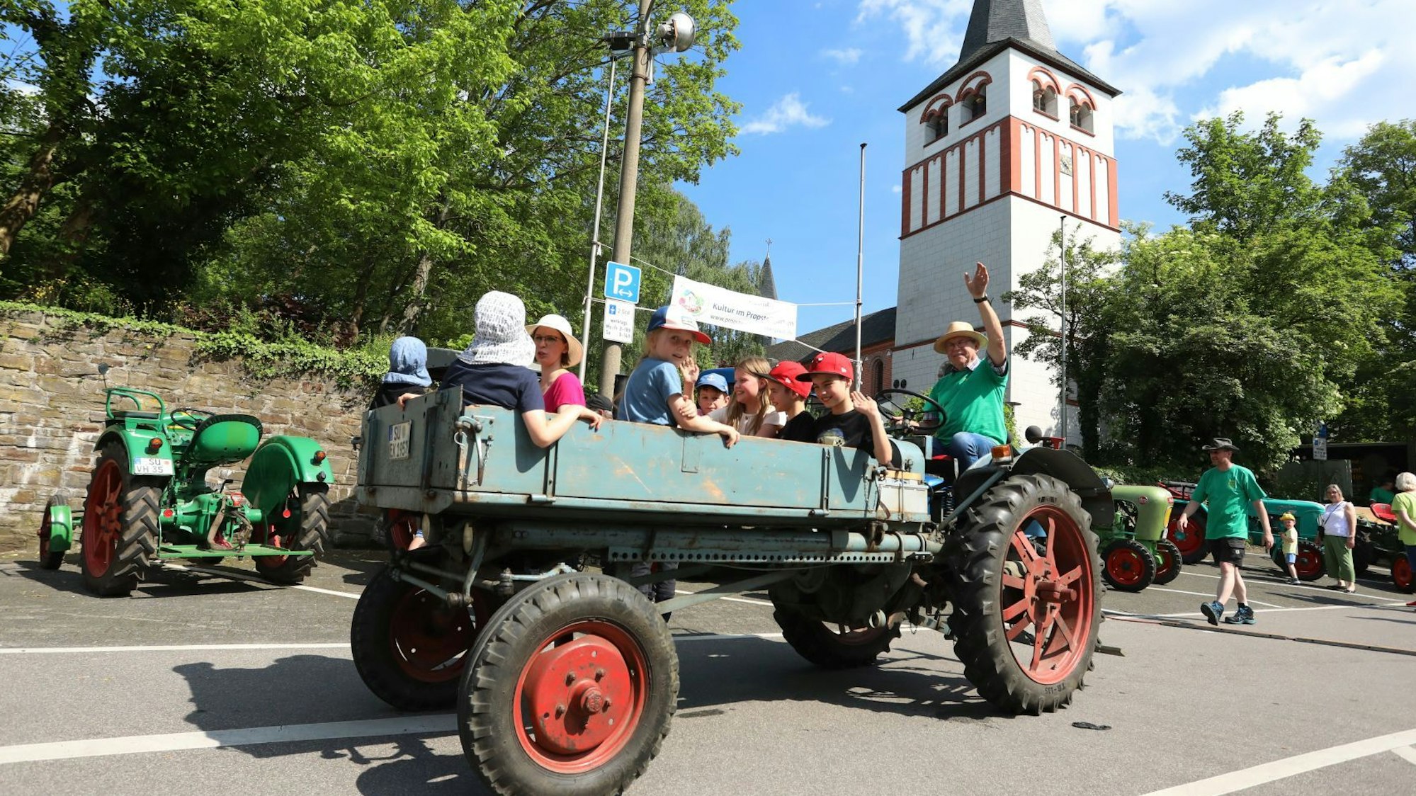 Auf der Ladefläche eines alten Traktors werden Kinder und Erwachsene durch den Ort gefahren, im Hintergrund steht ein rot-weißer Kirchturm.