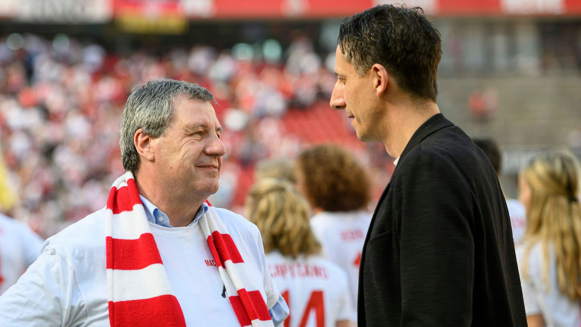 FC-Präsident Dr. Werner Wolf mit Vereinsschal und FC-Geschäftsführer Christian Keller unterhalten sich im Stadion; im Hintergrund ist undeutlich eine besetzte Stadiontribüne zu sehen.