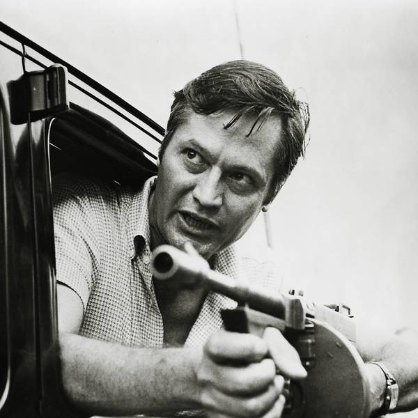 Roger Corman lehnt sich mit einer Maschinenpistole in den Händen aus einem Autofenster.