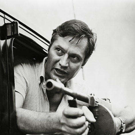 Roger Corman lehnt sich mit einer Maschinenpistole in den Händen aus einem Autofenster.