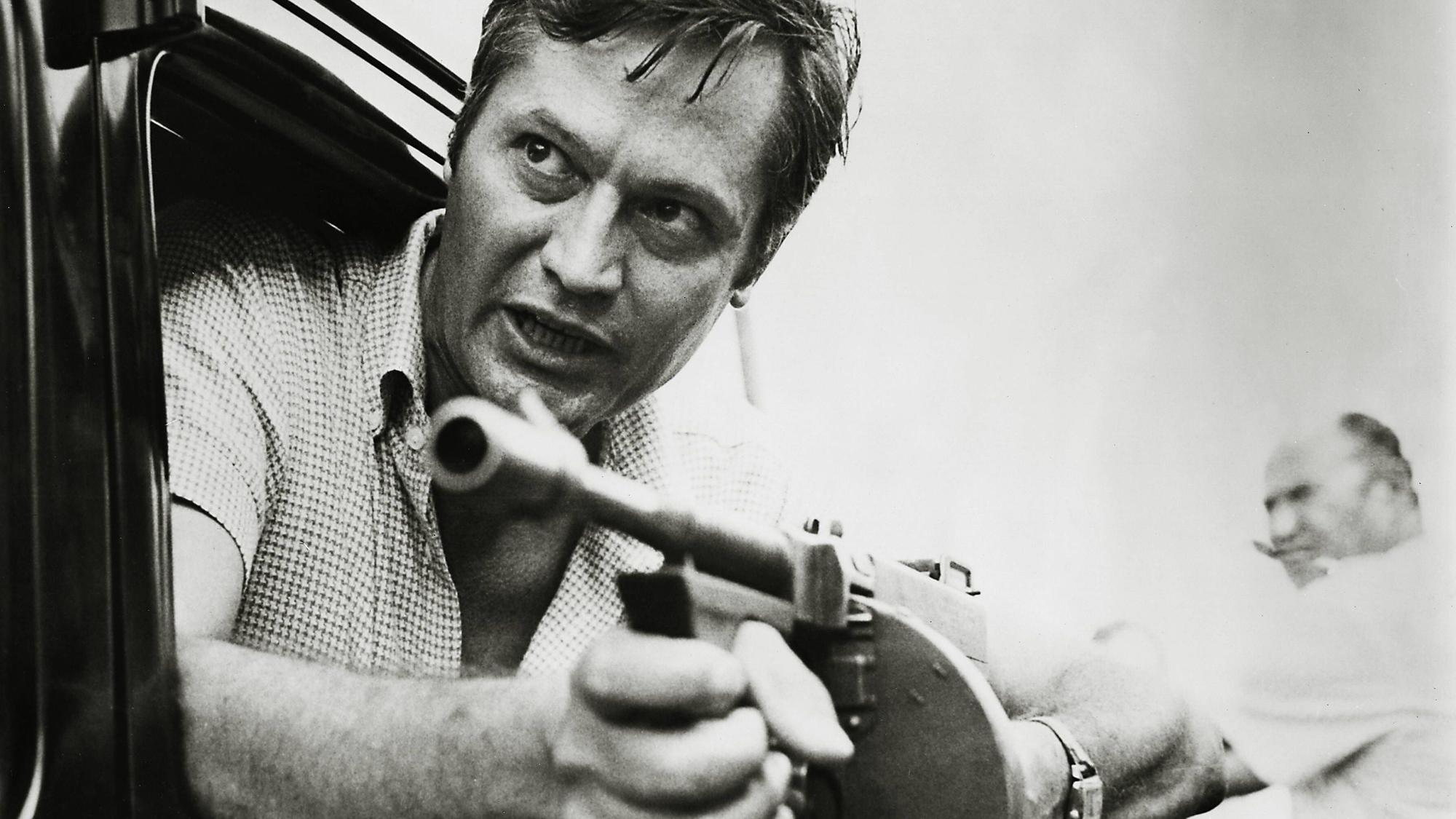 Roger Corman lehnt sich mit einer Maschinenpistole in den Händen aus einem Autofenster.