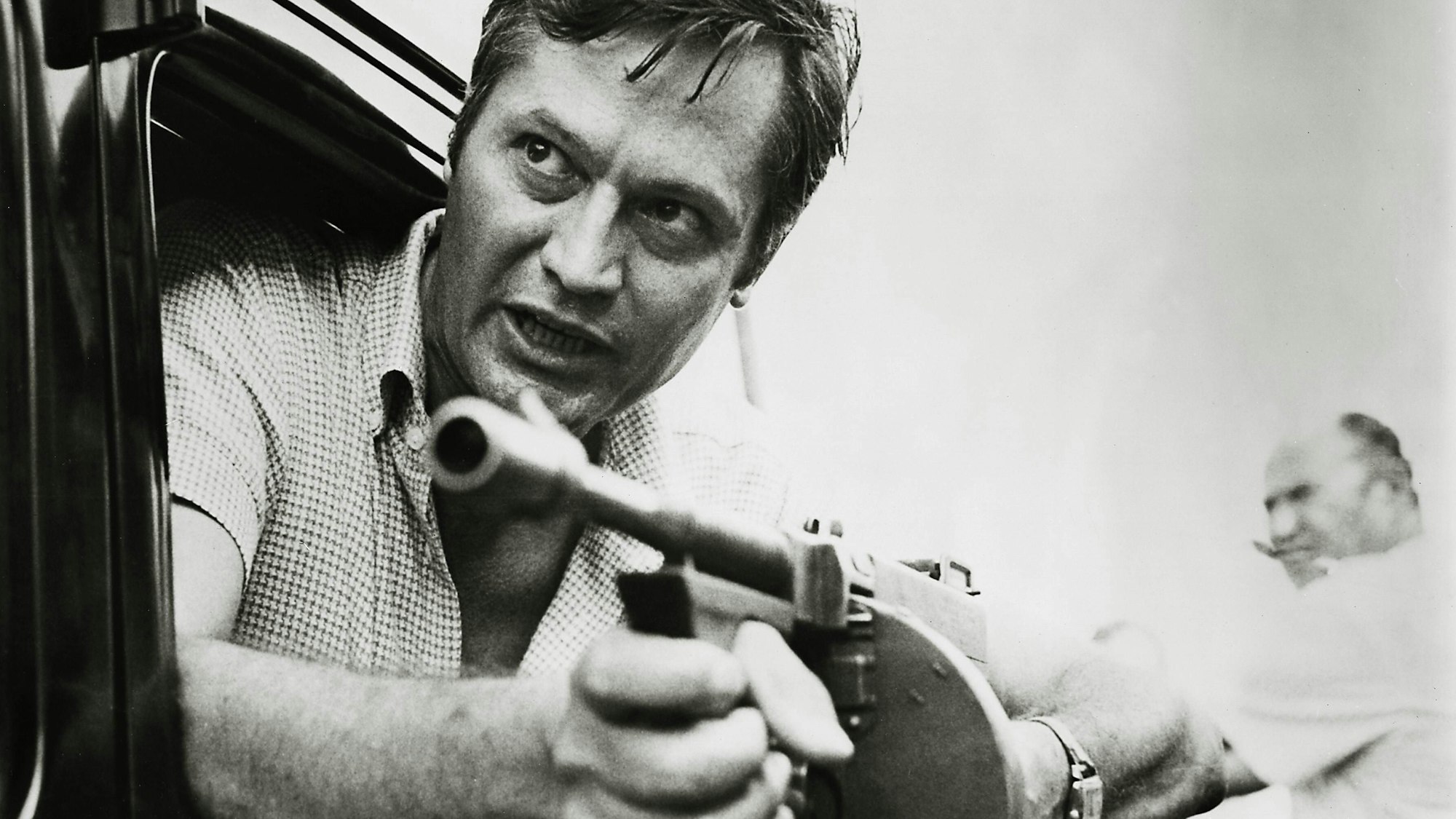 Roger Corman lehnt sich mit einer Maschinenpistole in den Händen aus einem Autofenster.