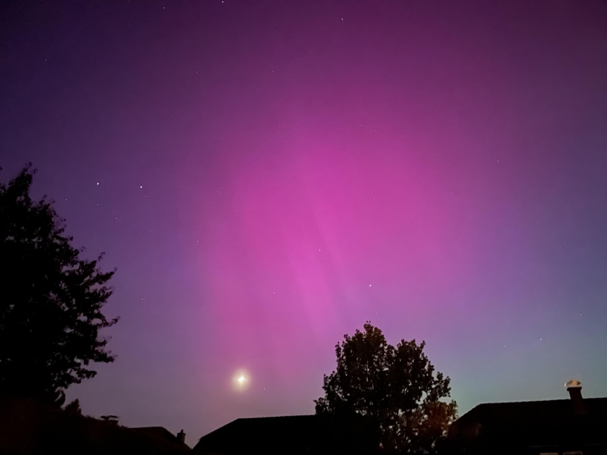 Über Hausdächern und Bäumen sieht man den Mond und ein Polarlicht.