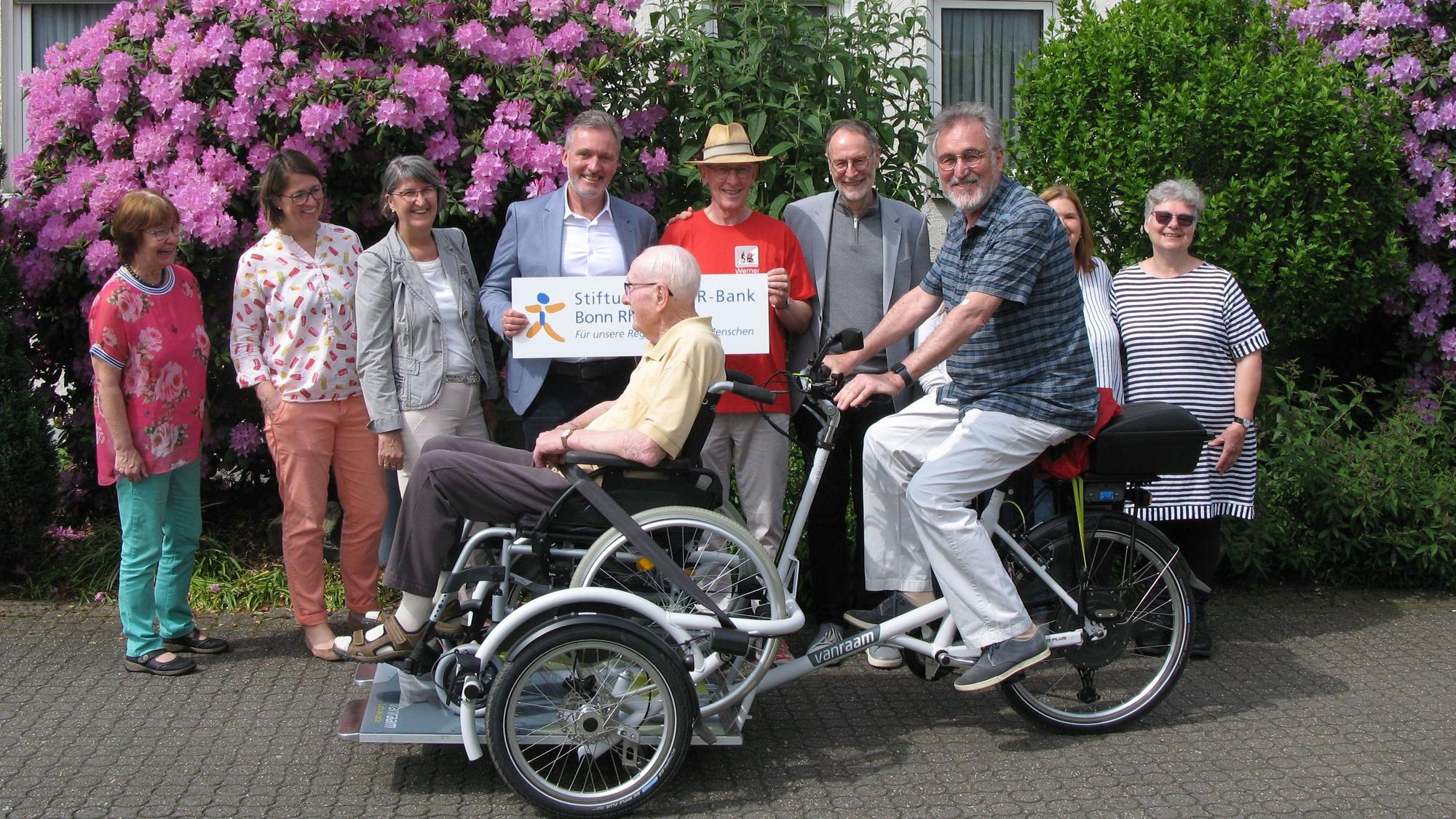 Die Rikscha mit Rollstuhlplattform, auf der ein Senior im Rollstuhl sitzt, fährt an eine Reihe von Menschen vorbei.
