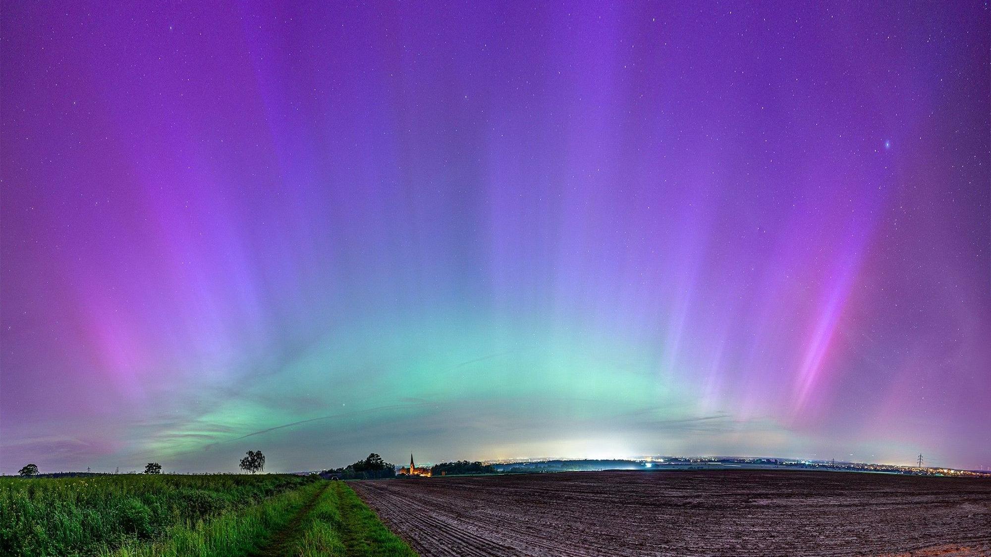 Über einem Feld und einem Kirchturm schillert der Himmel grün und violett.