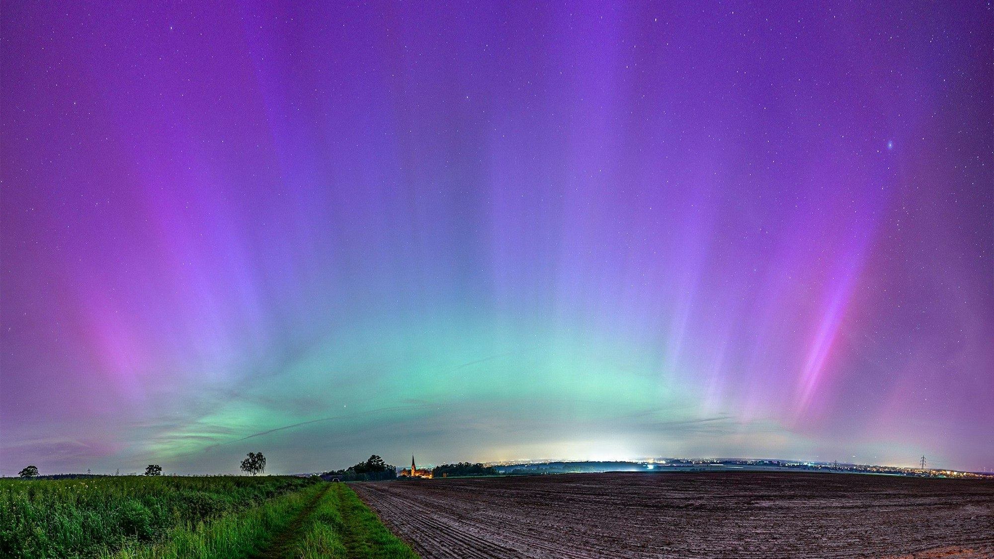 Über einem Feld und einem Kirchturm schillert der Himmel grün und violett.