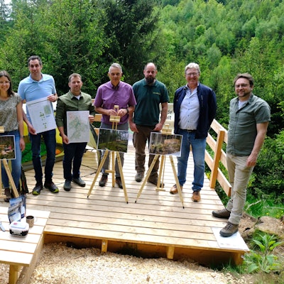 Vertreter der beteiligten Kommunen, des Naturparks Nordeifel und der Nordeifel Tourismus GmbH stehen auf einer Holzplattform im Wald.