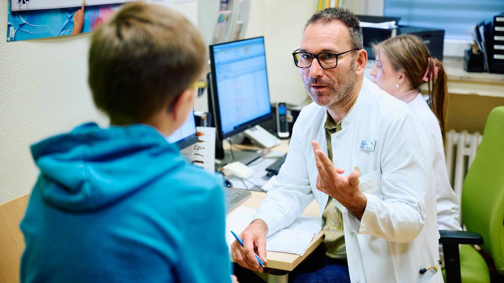 Kinder-Gastroenterologe Jens Berrang unterhält sich im Arztzimmer mit Zöliakie-Patient Jan Jonathan. Der Junge sitzt ihm gegenüber auf einer Liege, ist nur von hinten abgebildet und trägt einen blauen Kapuzenpullover.