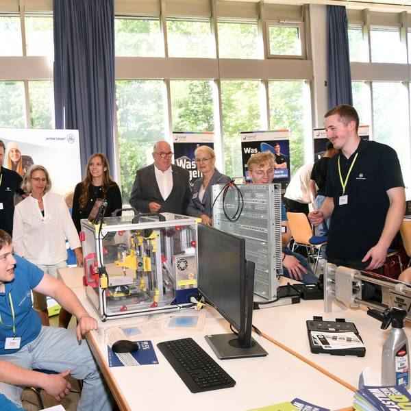 Junge Leute arbeiten an einer technischen Apparatur, Lehrer und Offizielle stehen dahinter.