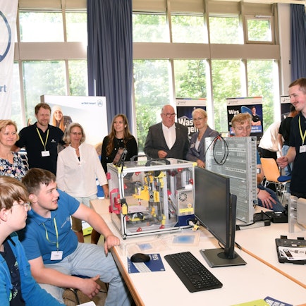 Junge Leute arbeiten an einer technischen Apparatur, Lehrer und Offizielle stehen dahinter.