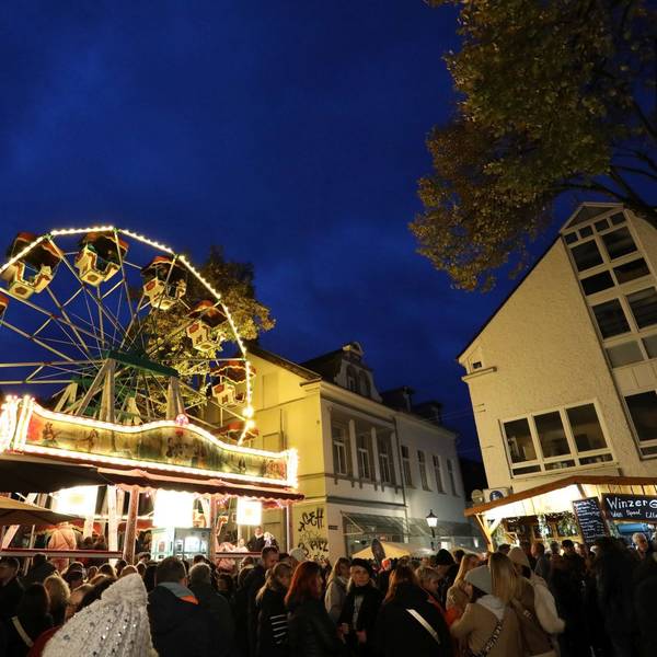Menschen tummeln sich auf einem Platz, auf dem mehrere Buden und ein beleuchtetes Riesenrad stehen.