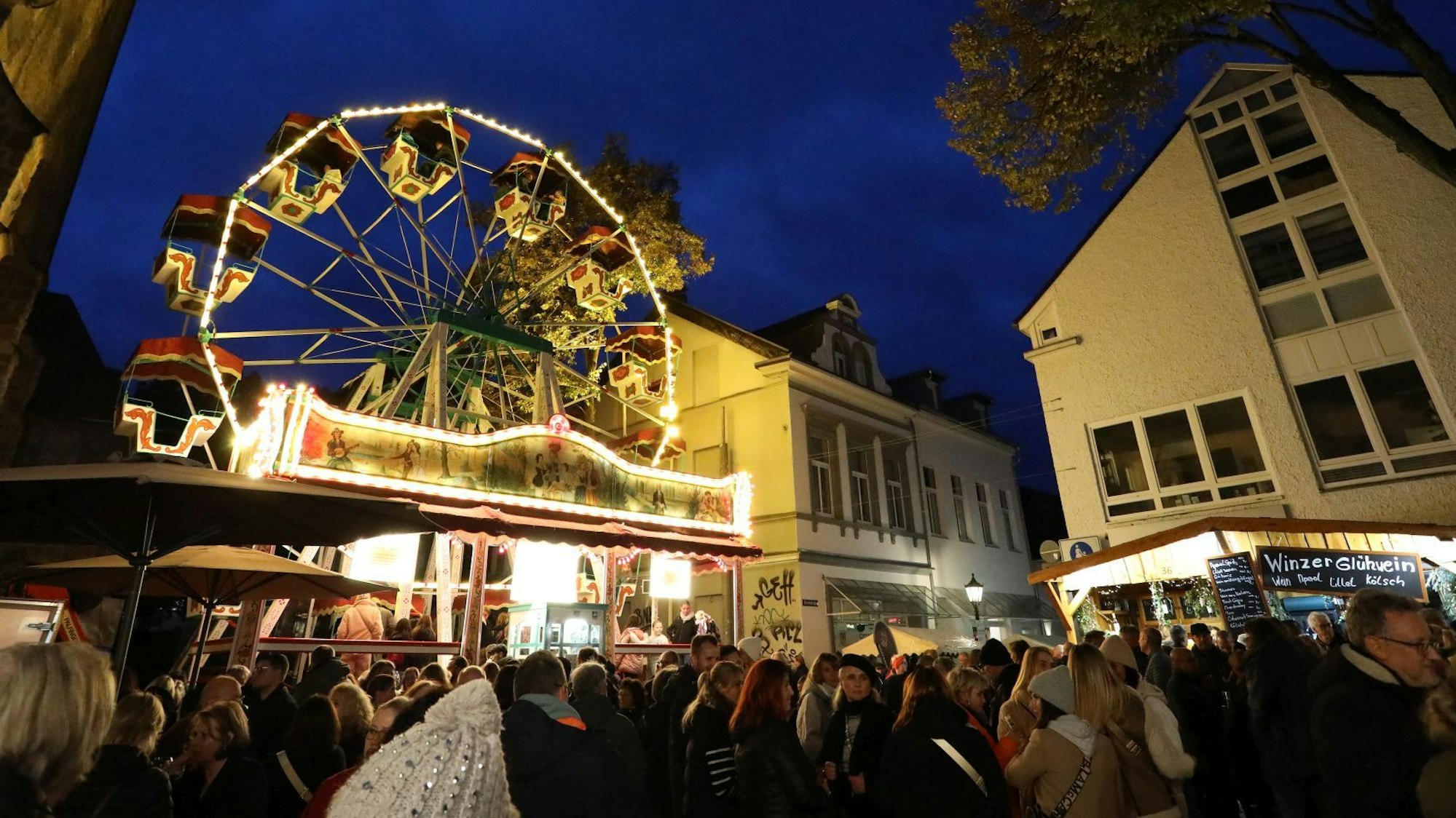 Menschen tummeln sich auf einem Platz, auf dem mehrere Buden und ein beleuchtetes Riesenrad stehen.