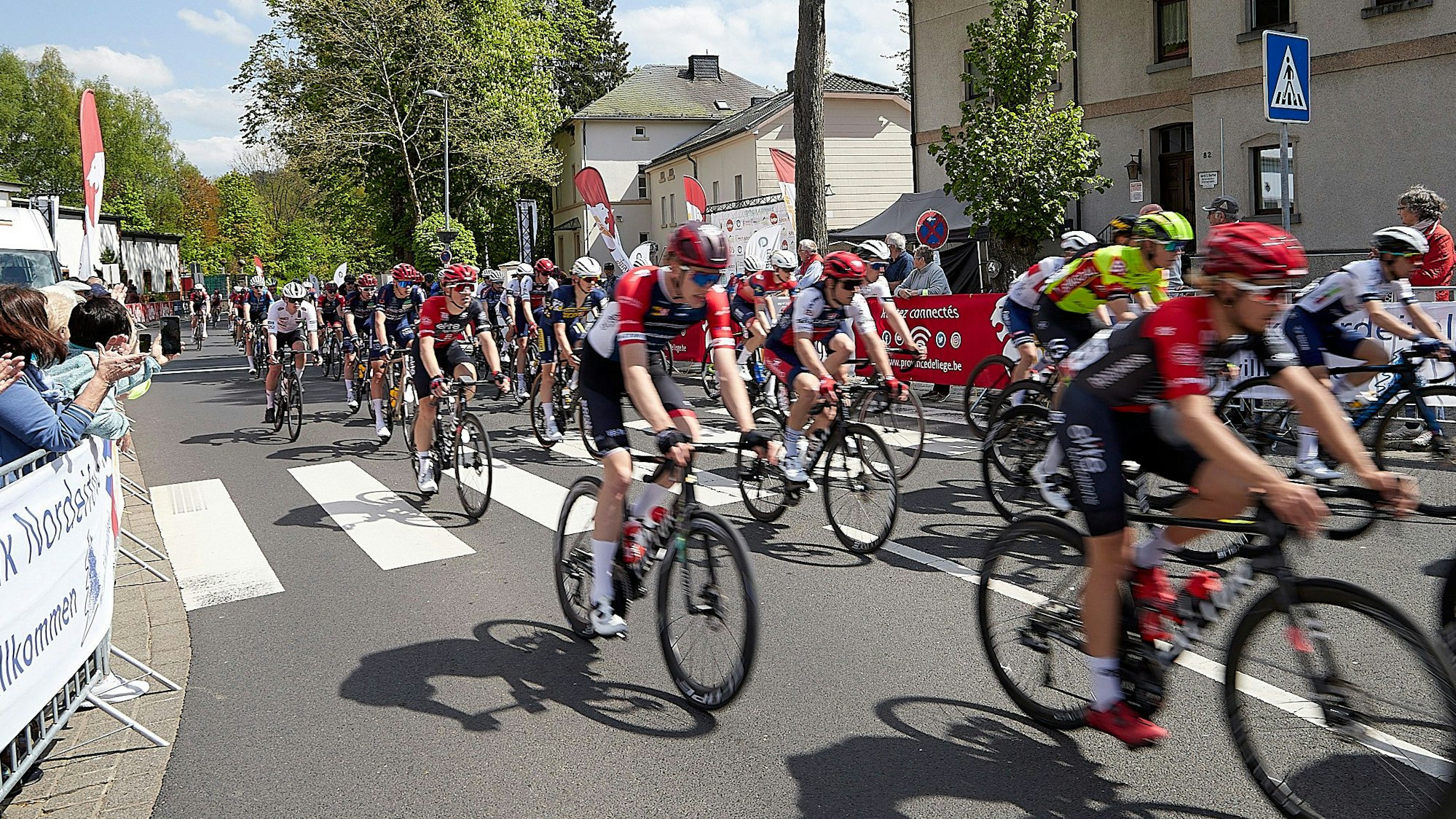 Zahlreiche Radrennfahrer fahren durch den Ortskern von Hellenthal. Hinter den Absperrungen stehen einige Zuschauer und applaudieren.