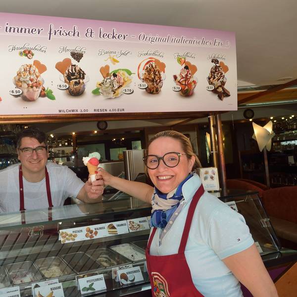 Ein Mann reicht einer Frau eine Eiswaffel mit drei Kugeln über die Theke. Beide Personen lächeln in Richtung der Kamera.