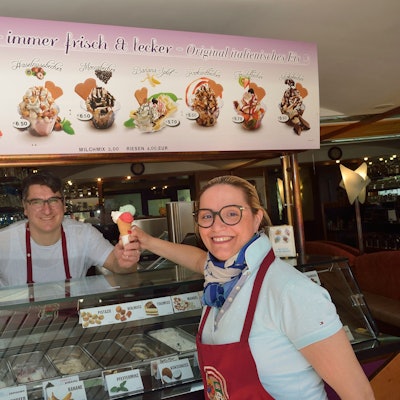 Ein Mann reicht einer Frau eine Eiswaffel mit drei Kugeln über die Theke. Beide Personen lächeln in Richtung der Kamera.