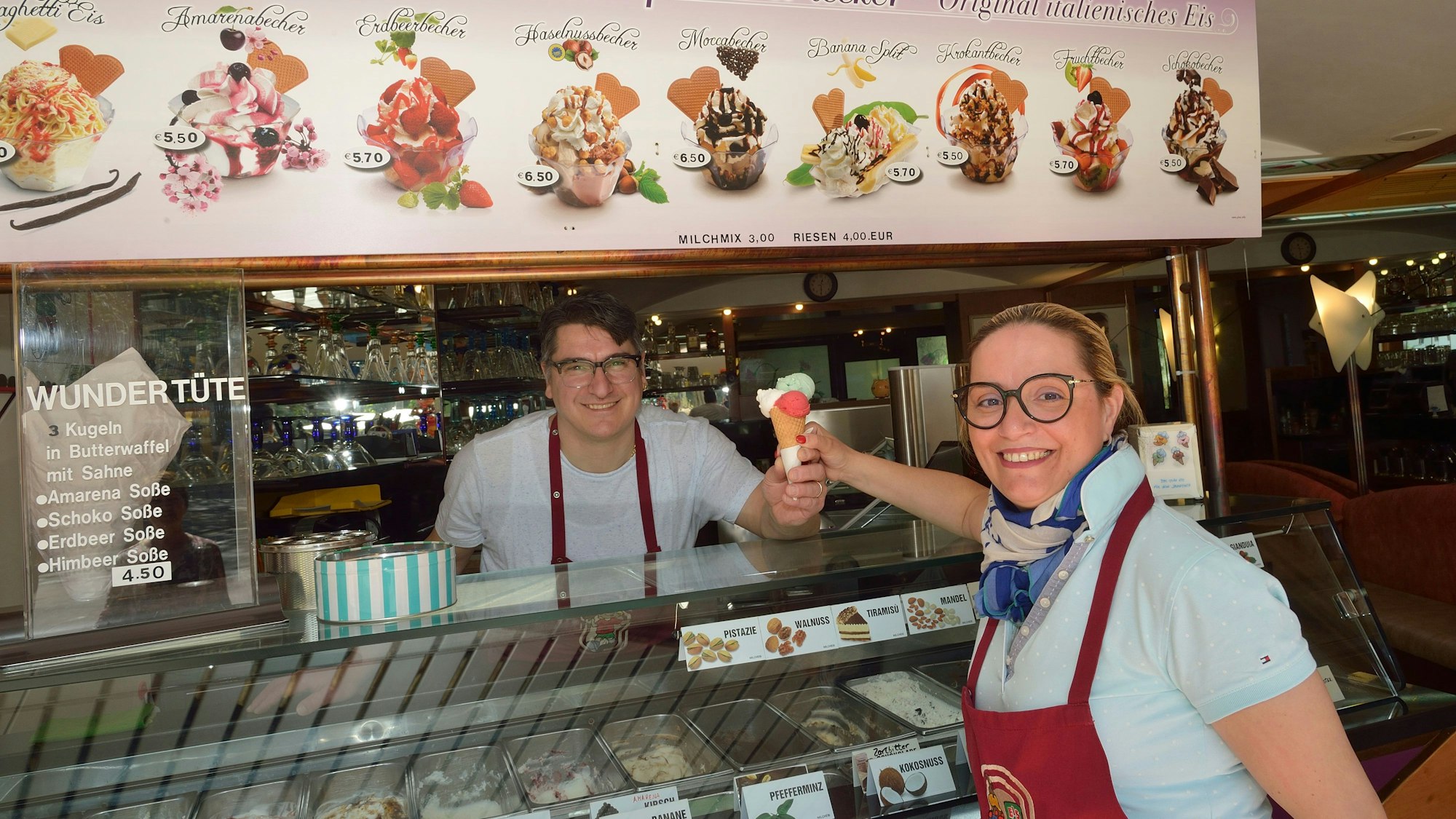 Ein Mann reicht einer Frau eine Eiswaffel mit drei Kugeln über die Theke. Beide Personen lächeln in Richtung der Kamera.