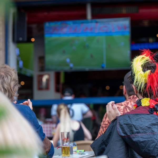 Ein Fan mit Deutschland-Perücke schaut ein Fußballspiel auf einer Leinwand.