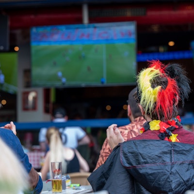 Ein Fan mit Deutschland-Perücke schaut ein Fußballspiel auf einer Leinwand.