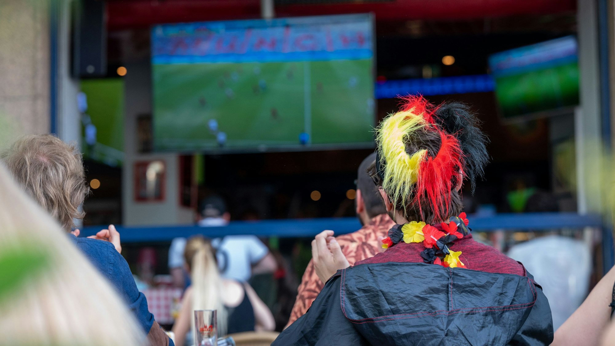 Ein Fan mit Deutschland-Perücke schaut ein Fußballspiel auf einer Leinwand.