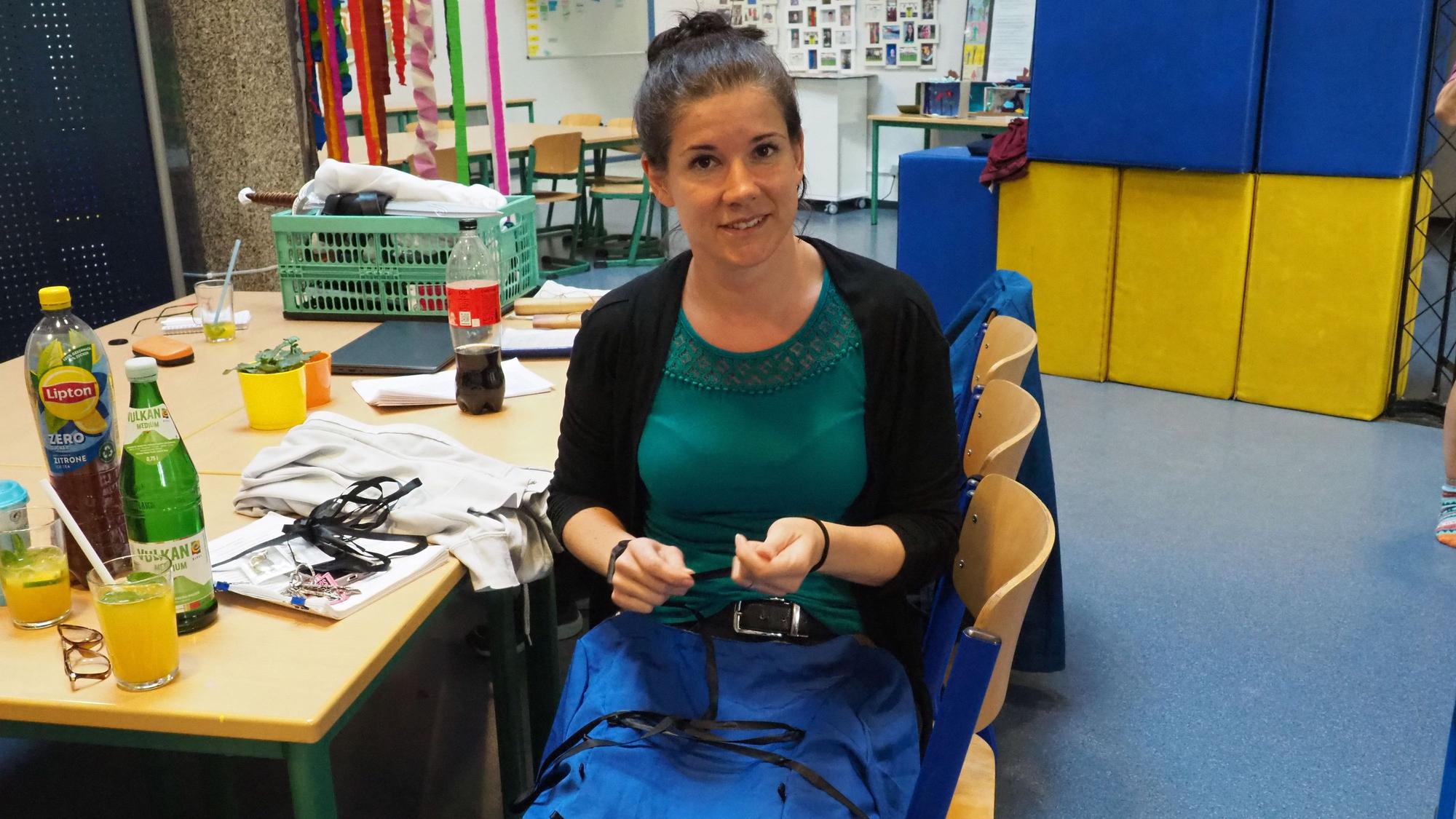 Susanna Janssen hat ihr Kostüm auf dem Schoß und arbeitet daran, im Hintergrund sind Requisiten zu sehen.