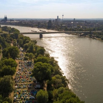 Schon während der Gamescom zelteten Besucher im Jugendpark: Ein Luftbild eines Zeltlagers auf der Landzunge am Rhein, im Hintergrund Kölns Skyline mit dem Dom.