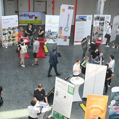 Das Foto zeigt Menschen, die eine Messe besuchen. Zu sehen sind auch Stehtische und Werbe-Aufsteller.