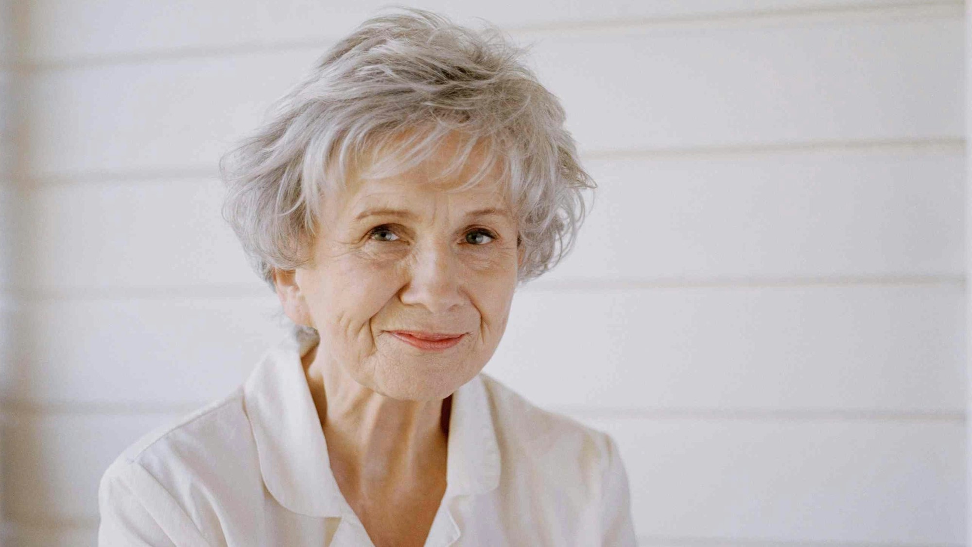 Alice Munro sitzt vor einer weißen Holzwand.