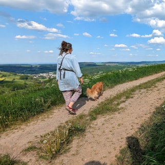 Eine Spaziergängerin und ihr Hund auf der Eifelspur „Soweit das Auge reicht“. Rechts an einem Zaunpfahl ist eine Wegmarkierung mit dem Titel des Weges angebracht.