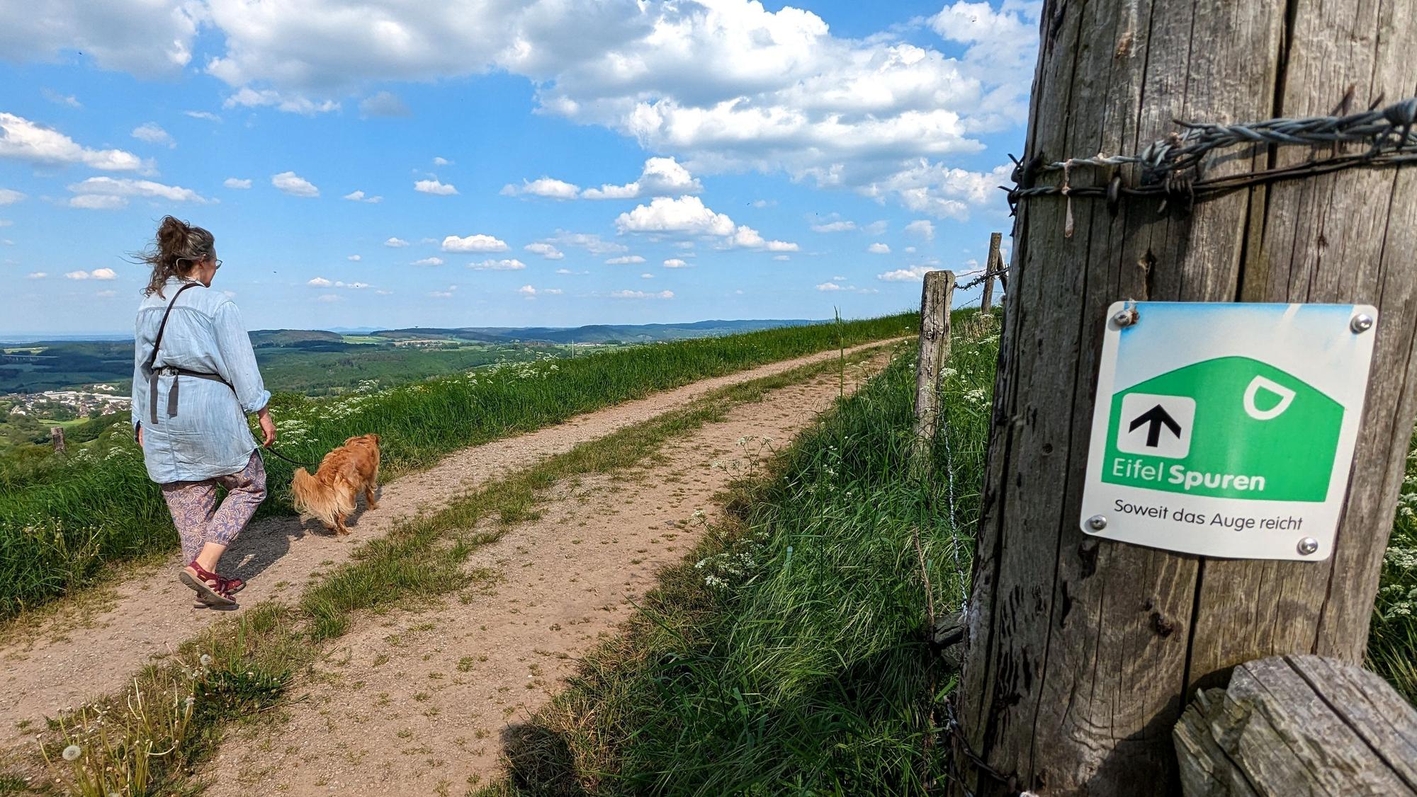 Eine Frau geht mit ihrem Hund auf einem Wanderweg. An einem Zaunpfahl ist eine EifelSpuren-Wegemarkierung angebracht.