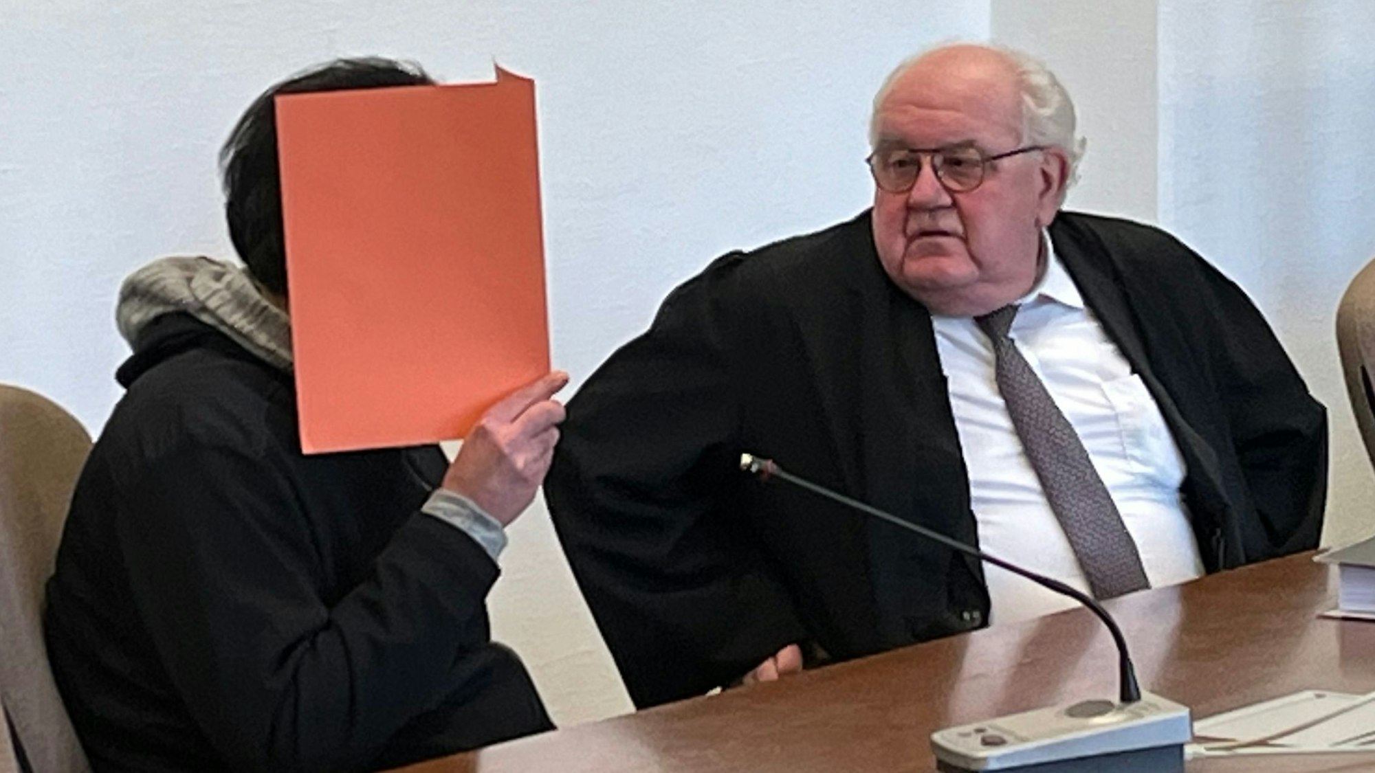 Der des Mordes angeklagte Rheindorfer sitzt, verborgen hinter einem Aktendeckel, neben seinem Verteidiger Gottfried Reims.