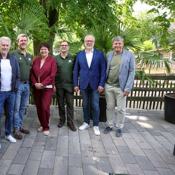 Sieben Menschen stehen auf einer Terrasse vor Bäumen und schauen in die Kamera.