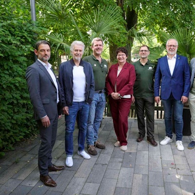 Sieben Menschen stehen auf einer Terrasse vor Bäumen und schauen in die Kamera.