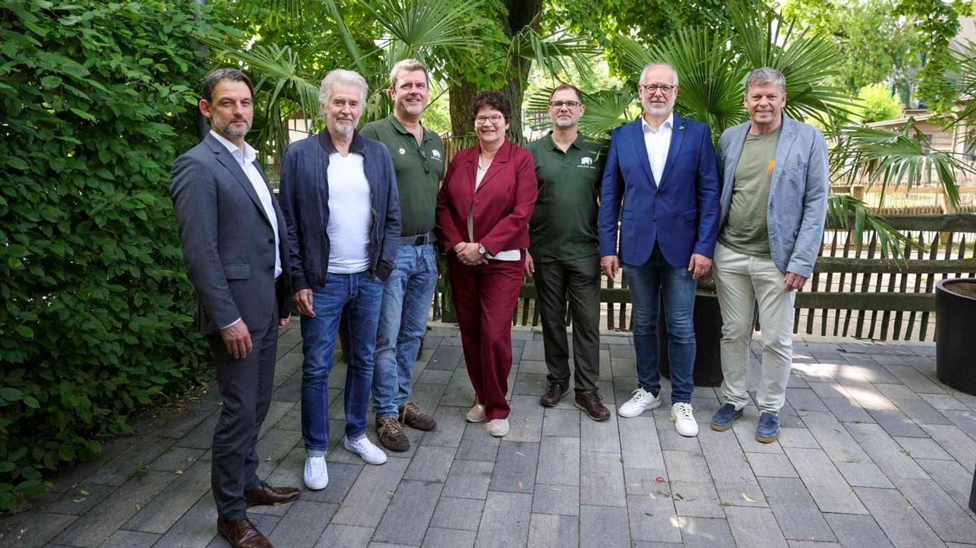 Sieben Menschen stehen auf einer Terrasse vor Bäumen und schauen in die Kamera.
