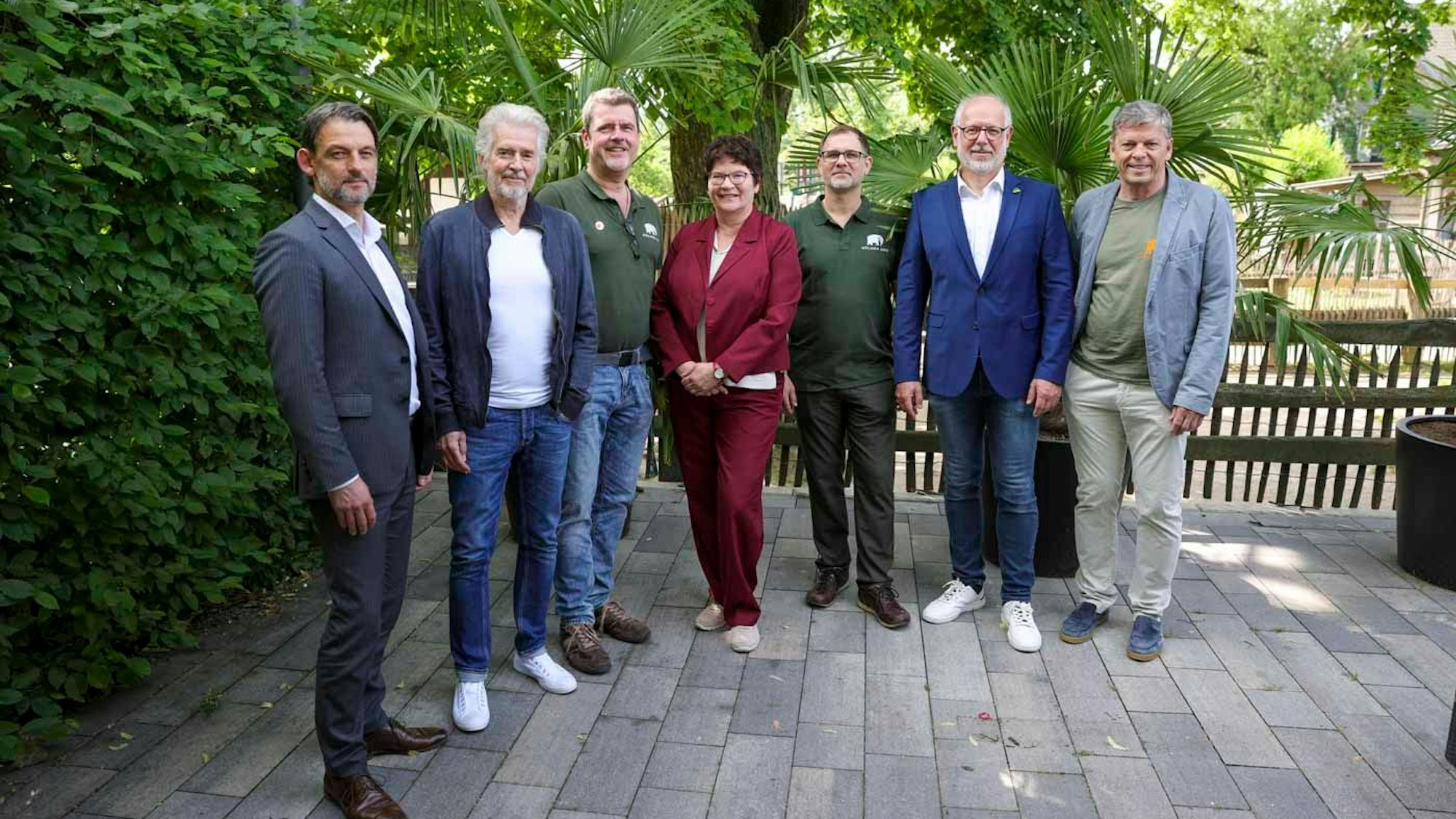 Sieben Menschen stehen auf einer Terrasse vor Bäumen und schauen in die Kamera.