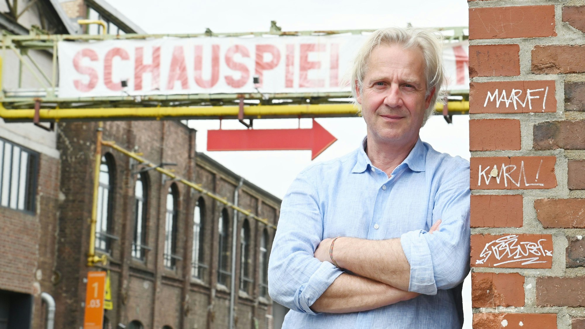 15.05.2024 Köln. Stefan Bachmann lehnt sich an die Ecke des Depot 2 in Köln-Mülheim. Hinter ihm das Die Straße überspannende "Schauspiel Köln"-Schild. Der rote Pfeil, der zum Eingang weist, scheint direkt auf ihn zu zielen.