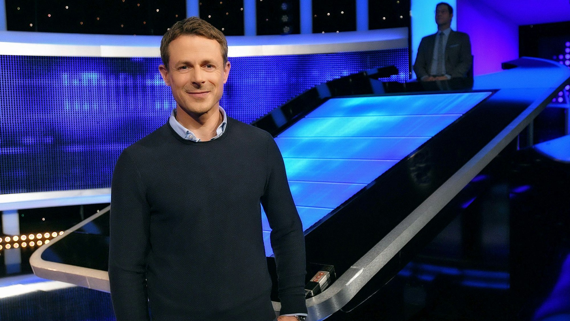 Moderator Alexander Bommes steht im Studio der ARD-Quizshow „Gefragt – Gejagt“ vor dem Sitz des Jäger-Teams, das die Kandidatinnen und Kandidaten vom Gewinn des Geldes abhalten soll.
