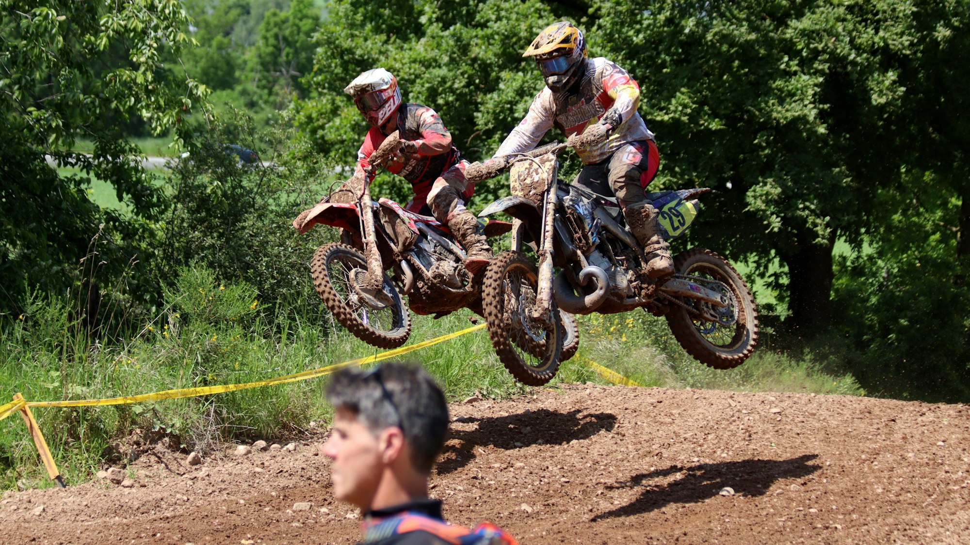 Zwei Motocross-Maschinen befinden sich samt ihrer Fahrer ausgangs eines Hügels in der Luft, beide sind voller Matsch. Im Vordergrund steht ein Zuschauer oder Streckenposten.
