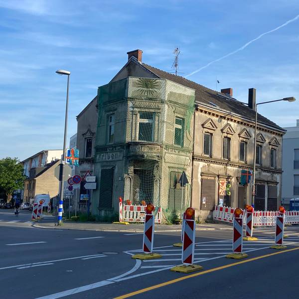 An einer zweispurigen Straße stehen viele rot-weiße Baken, ein Haus ist zum Teil von einem Schutznetz umgeben.