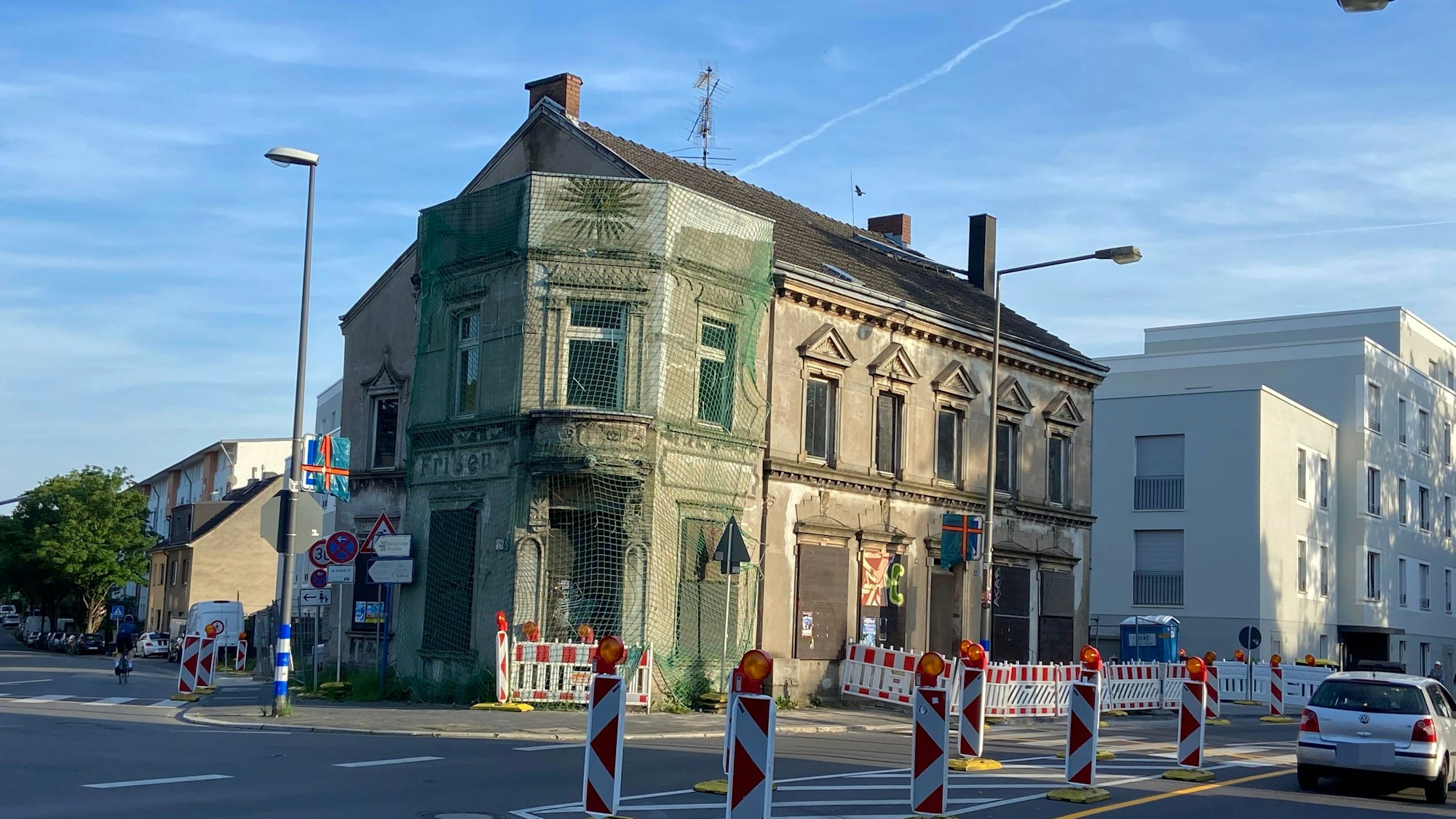 An einer zweispurigen Straße stehen viele rot-weiße Baken, ein Haus ist zum Teil von einem Schutznetz umgeben.