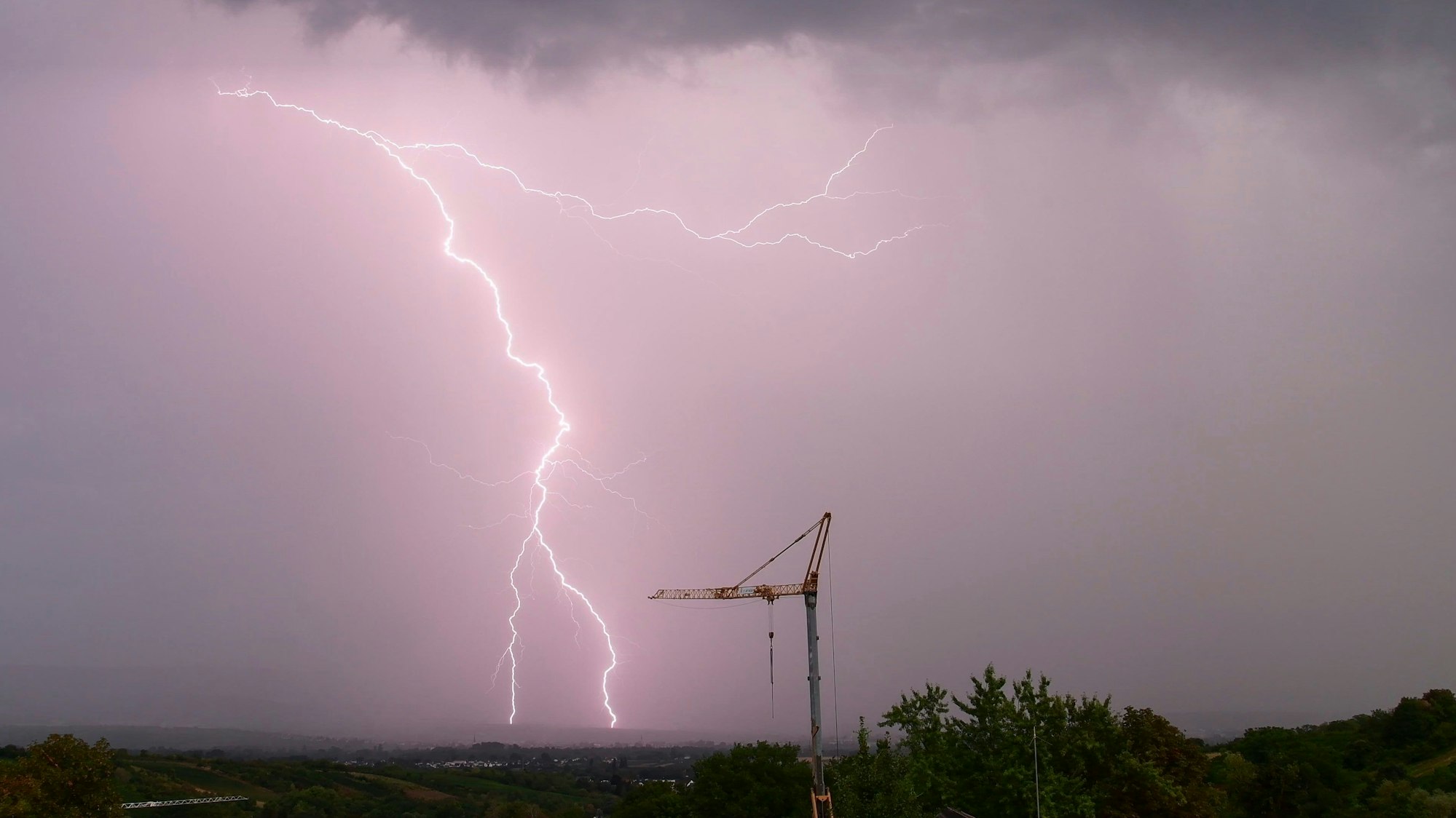 Ein gewaltiger Blitz schlägt am vom Unwetter lila gefärbten Himmel in der Nähe eines Baukrans ein. Schwere Unwetter werden in der Nacht zu Freitag in Köln und der Region erwartet. (Archivbild)