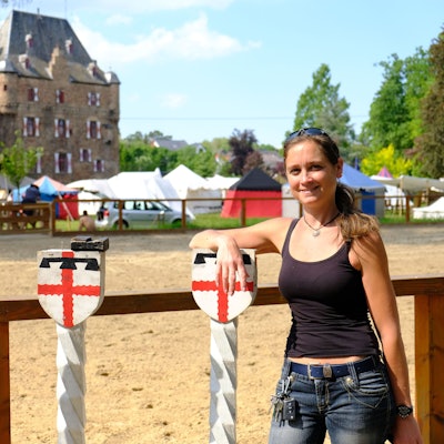 Patricia Gräfin Beissel steht mit einem schwarzen Top am Reitplatz vor Burg Satzvey, die im Hintergrund zu sehen ist.