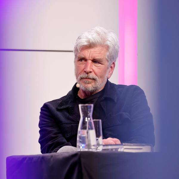 Karl Ove Knausgård sitzt auf dem Podium im großen Saal der Kölner Flora. Seine Haare und sein Vollbart sind weiß. Er trägt ein dunkelblaues Hemd.