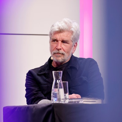 Karl Ove Knausgård sitzt auf dem Podium im großen Saal der Kölner Flora. Seine Haare und sein Vollbart sind weiß. Er trägt ein dunkelblaues Hemd.