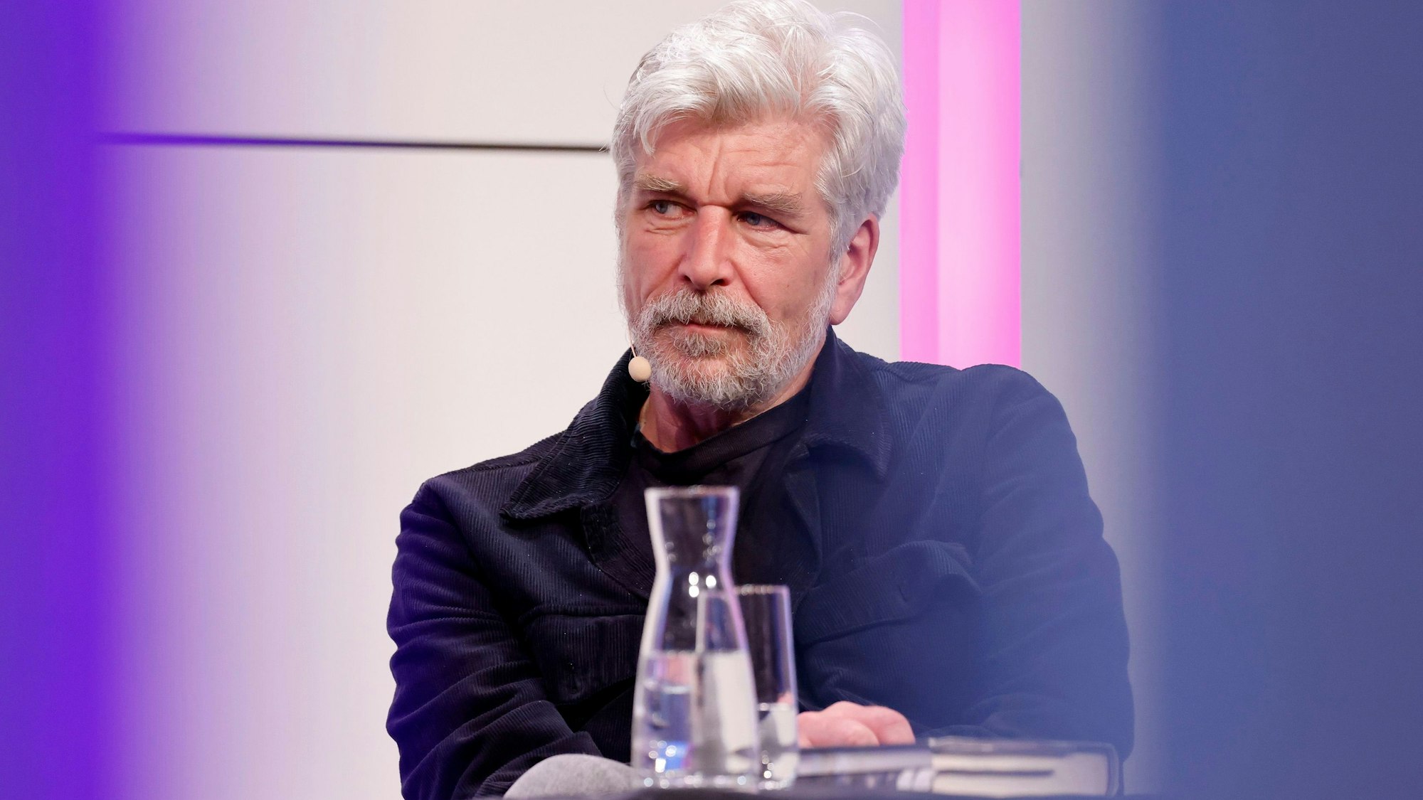 Karl Ove Knausgård sitzt auf dem Podium im großen Saal der Kölner Flora. Seine Haare und sein Vollbart sind weiß. Er trägt ein dunkelblaues Hemd.