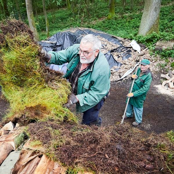 Gerd Linden bedeckt die gestapelten Scheite mit Grassoden, ein Helfer schaut von unten zu.