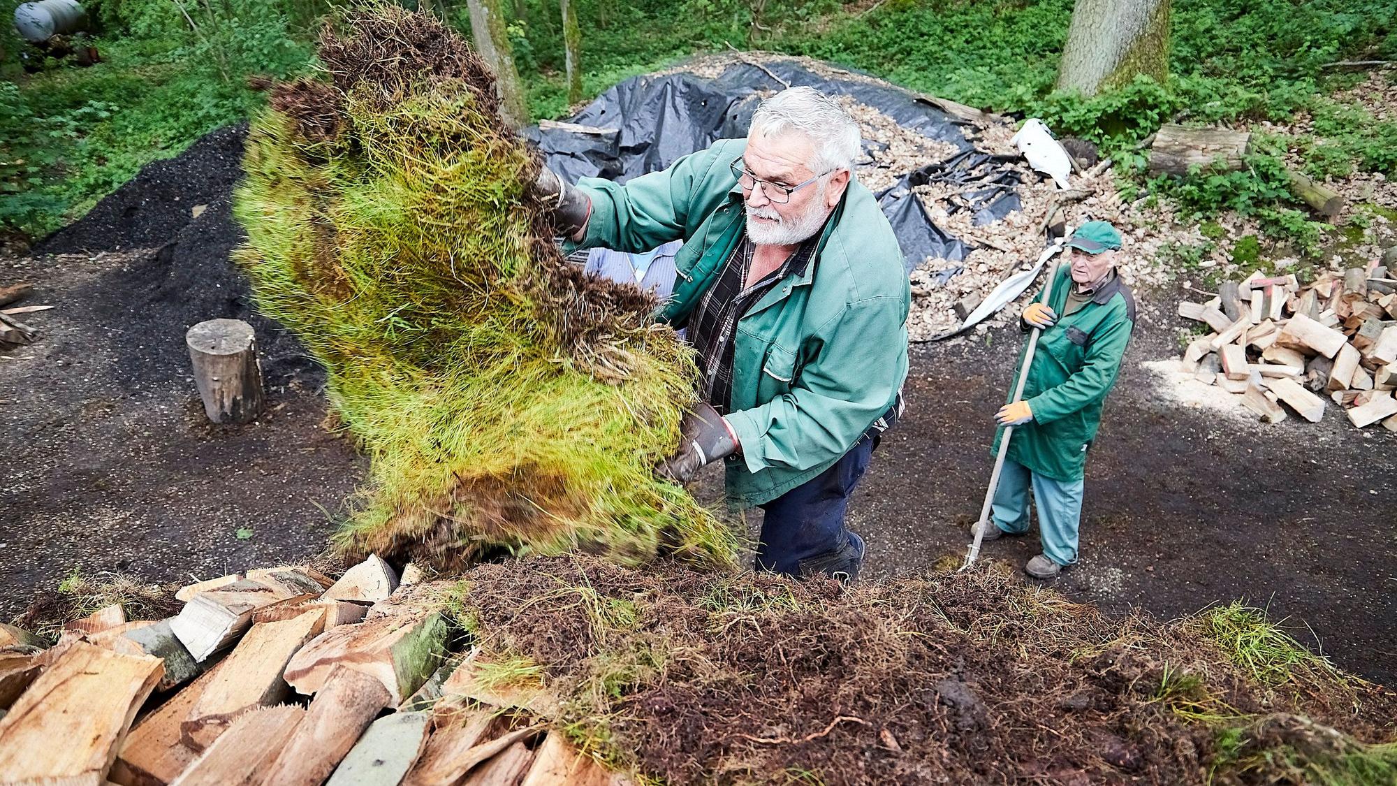 Gerd Linden bedeckt die gestapelten Scheite mit Grassoden, ein Helfer schaut von unten zu.