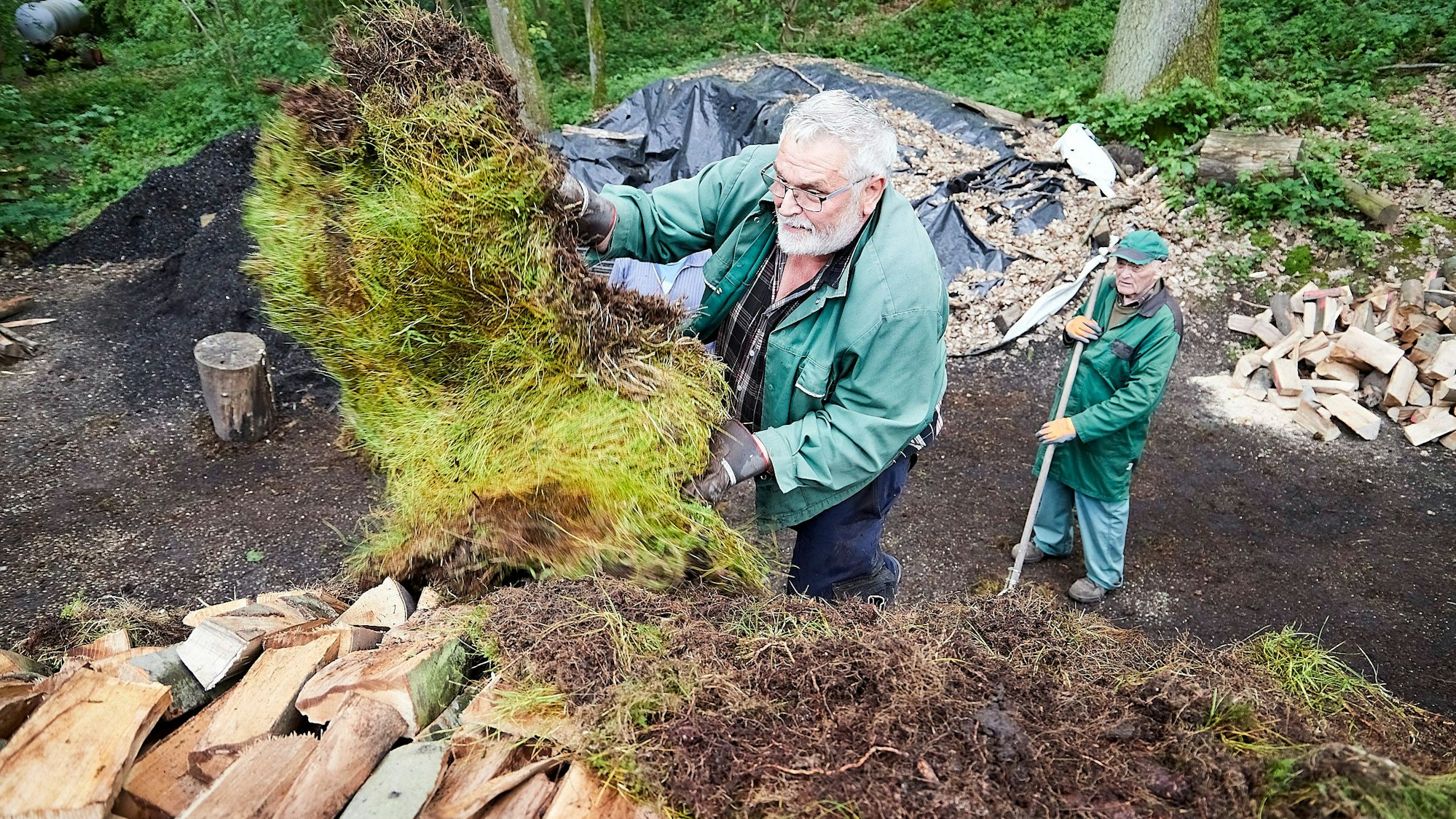 Gerd Linden bedeckt die gestapelten Scheite mit Grassoden, ein Helfer schaut von unten zu.