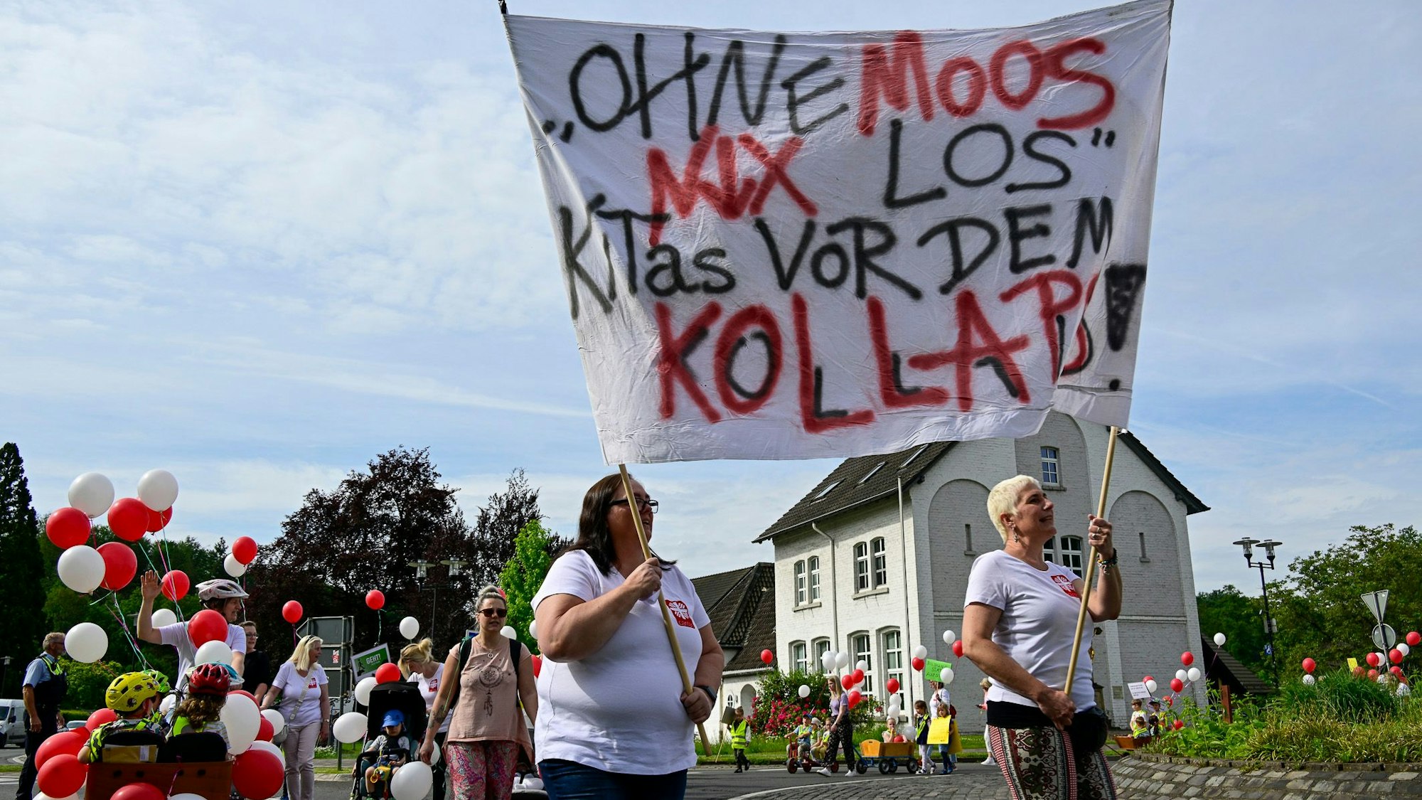 Zwei Erwachsene tragen ein großes Plakat mit der Aufschrift „Ohne Moos nix los“, dahinter folgen die anderen Demonstranten, darunter sind auch Kinder.