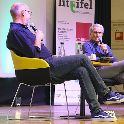 Zwei Männer sitzen auf einer Bühne und unterhalten sich.